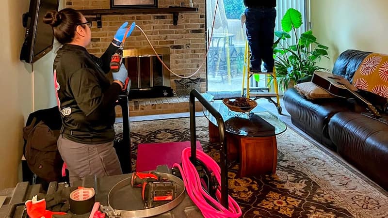 Two Luxe Ducts technicians cleaning a living room air duct, one on a step ladder at the ceiling register and one at the equipment cases on the floor