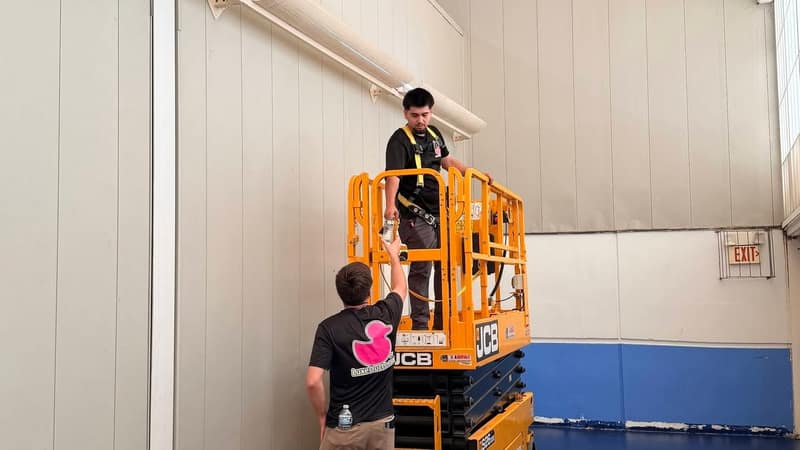 A Luxe Ducts technician on a scissor lift reaching a high wall vent in a gymnasium while a second technician spots from the floor