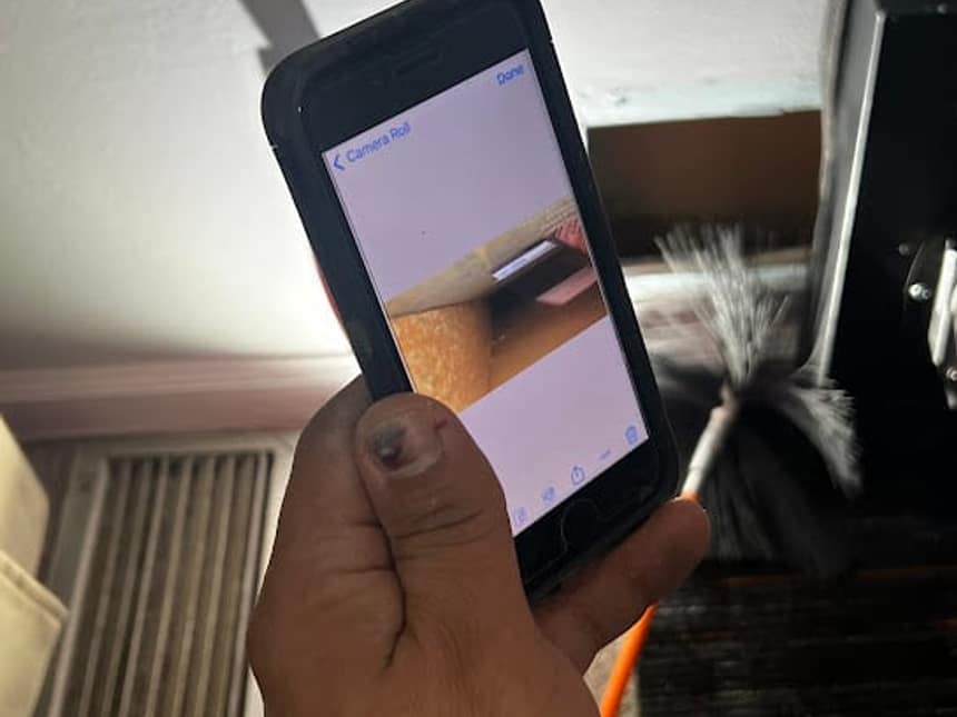 A technician holding a phone up to a wall register, photographing the inside of the duct with the cleaning brush still in the opening